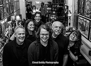 Seven band members posing and smiling at the camera with a wall of photos on both sides of them.